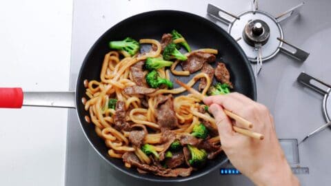 Stirring beef and broccoli yaki udon in a a large frying pan with chopsticks.