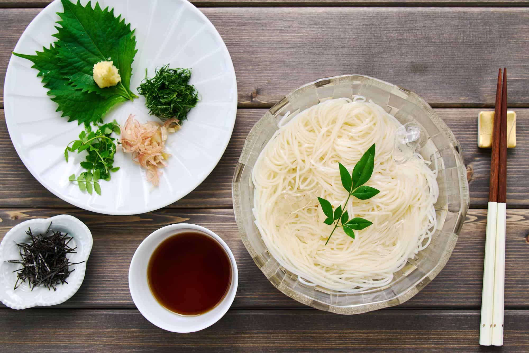 Overhead shot of a bowl filled with chilled somen noodles in ice water, poised next to a cup of flavorful dipping sauce and a plate of vibrant condiments.