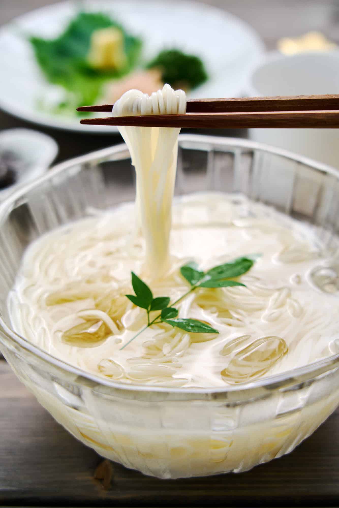 Close up of whisper thin strands of chilled somen noodles being lifted from a bowl of ice water set against a backdrop of vibrant condiments.