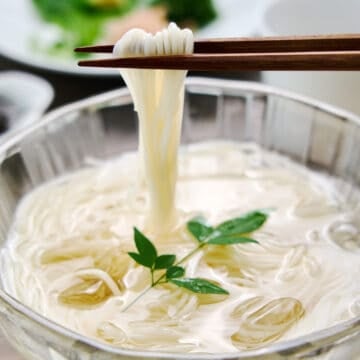 Close up of whisper thin strands of chilled somen noodles being lifted from a bowl of ice water set against a backdrop of vibrant condiments.