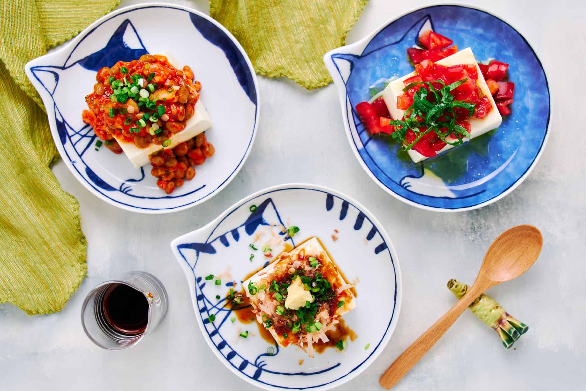 Overhead shot of Japanese cold tofu salad variations showcasing vibrant garnishes including a traditional hiyayakko at the bottom, a kimchi and natto one at the top left and a Caprese Salad variation at the top right.