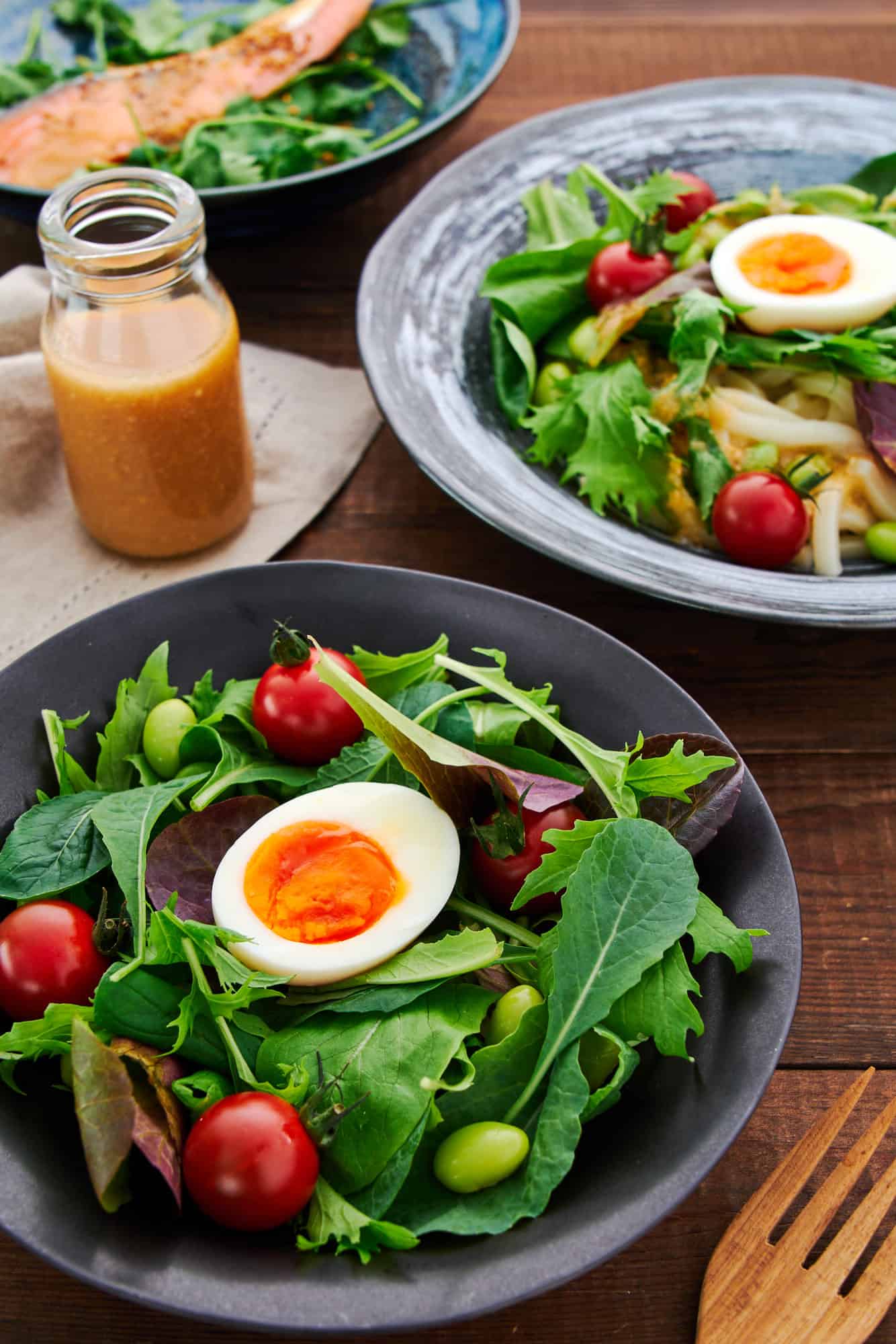 High-angle shot showing a vibrant table spread with a baby green salad, poached salmon, and cold udon salad, all enhanced by a bottle of Ginger Miso Dressing.