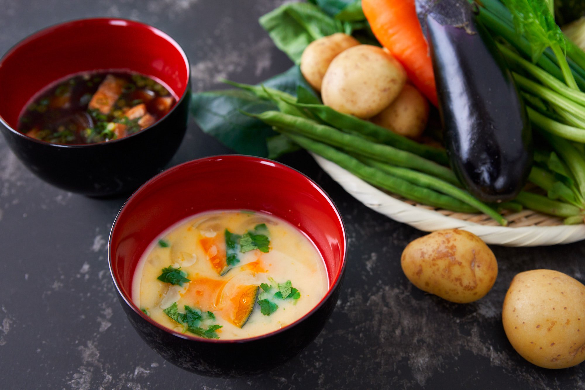 White miso soup with onions, kabocha, and mitsuba in the foreground with a red miso soup and basket of vegetables in the background.