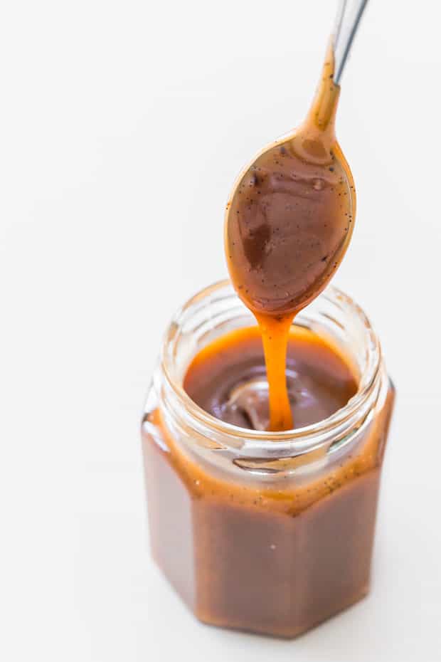 A jar of rich, delicious Vanilla Caramel Sauce. A ribbon of caramel pouring from a spoon into the jar.