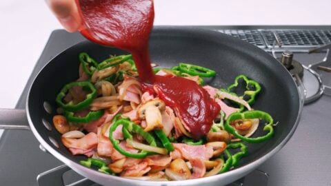 Adding ketchup to a pan with green peppers, bacon, mushrooms and onions for making Japanese spaghetti.