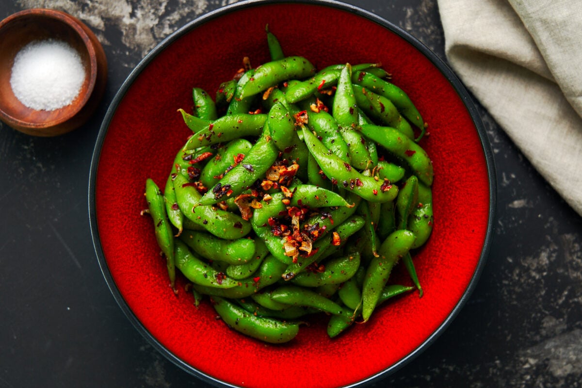 Edamame with garlic, black pepper, and chili peppers in a red bowl on a dark surface.