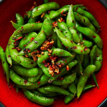 Edamame with garlic, black pepper, and chili peppers in a red bowl on a dark surface.