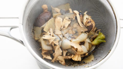 Mushrooms and charred aromatics in a strainer, straining plant-based pho broth.